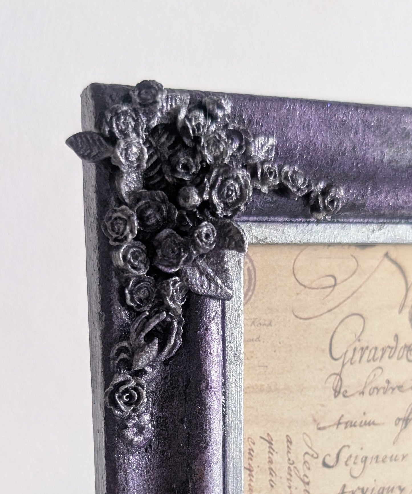 A close-up of a decorative picture frame with a distressed purple and black finish, featuring ornate floral corner embellishments and a vintage cursive script insert.