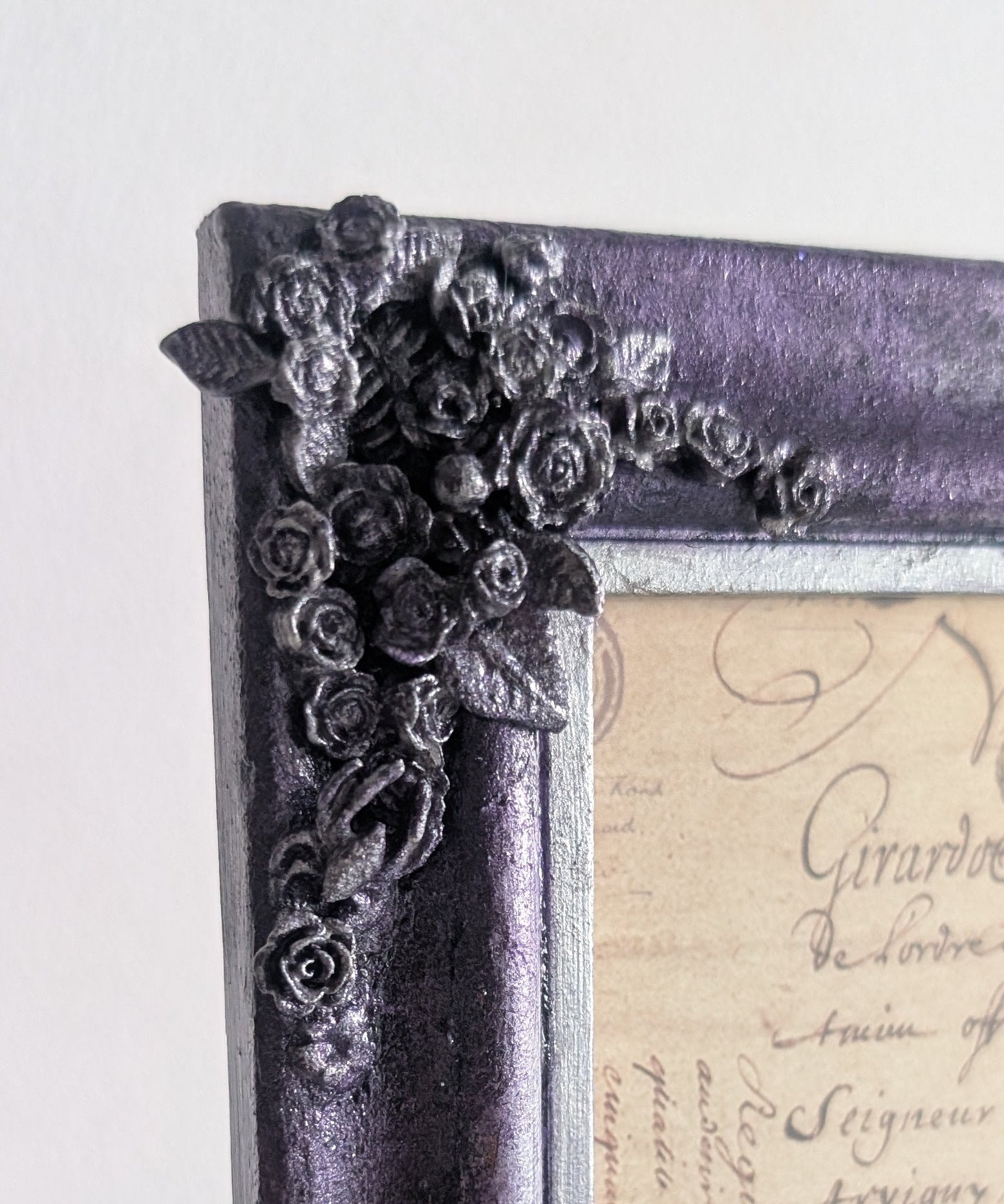 A close-up of a decorative picture frame with a distressed purple and black finish, featuring ornate floral corner embellishments and a vintage cursive script insert.