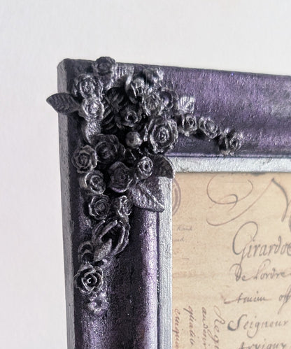 A close-up of a decorative picture frame with a distressed purple and black finish, featuring ornate floral corner embellishments and a vintage cursive script insert.