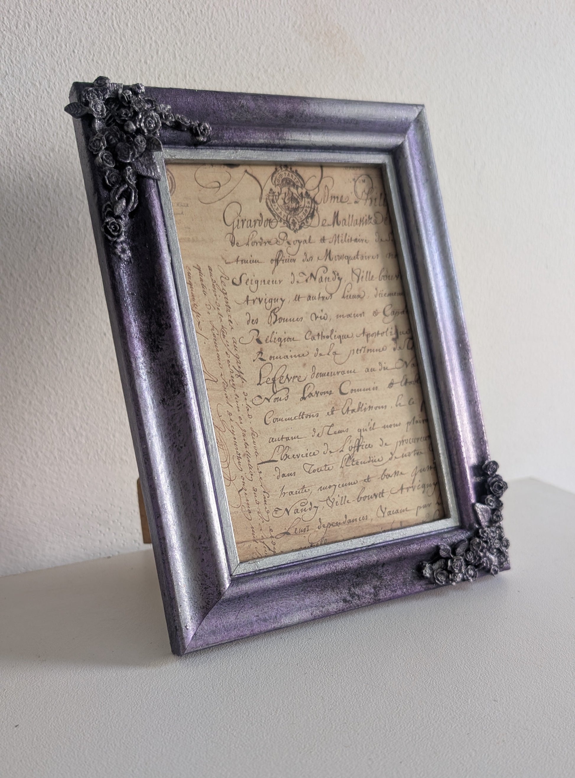 A vintage-style picture frame with ornate floral corner details in a distressed purple and silver finish, holding a piece of antique cursive handwriting on aged beige paper, shown standing on a light surface against a plain wall.