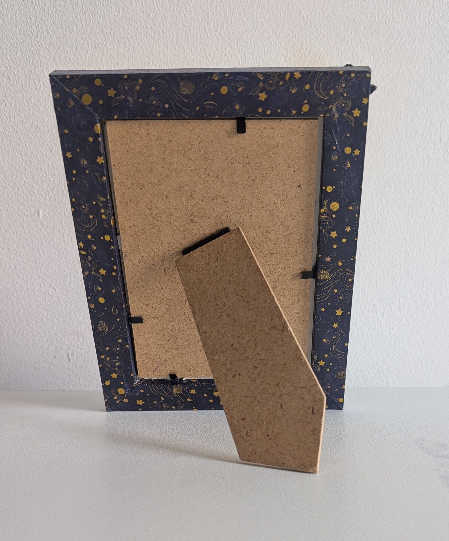 The back of a square picture frame with a speckled navy blue border and gold star details, standing on a white surface against a white wall.